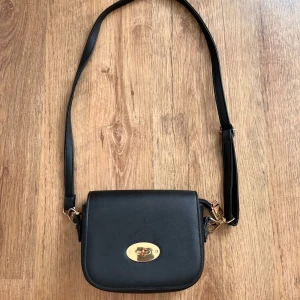 Svart axelväska med gulddetaljer - Säljer en stilren svart axelväska i syntetmaterial med snygga gulddetaljer. Väskan har en justerbar axelrem och ett elegant lås framtill. Perfekt för både vardag och fest!