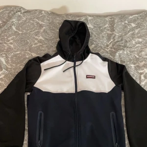 Svart och vit vindjacka från McKenzie - Säljer en snygg svart, mörkblå och vit zip hoodie från McKenzie. Tröjan har en dragkedja framtill och en huva. Perfekt för blåsiga dagar och har en sportig look. Den har också praktiska fickor med dragkedja.