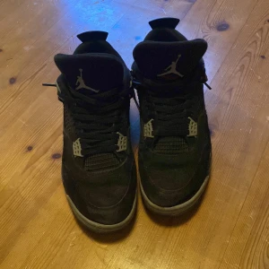 Svarta Jordan 4s - Säljer ett par svarta Nike Air Jordans med den ikoniska Jumpman-loggan. Skorna har en robust design med snörning och synlig Air-dämpning i sulan. Perfekta för sneakerheads som vill ha en klassisk stil. Pris är ej hugget i sten