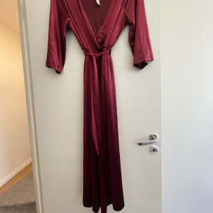 Vinröd långklänning i satin - Elegant vinröd långklänning i satin med omlott-design och knytband i midjan. Klänningen har trekvartsärmar och en v-ringad halslinje, perfekt för en stilfull look.
