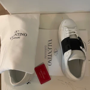 Valentino Garavani sneakers i vitt och svart - Snygga Valentino Garavani sneakers i vitt med en bred svart rem över snörningen. Skorna har en klassisk design med rund tå och är tillverkade i skinn. Perfekta för en stilren look.