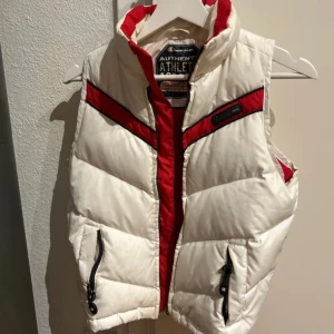 Vit och röd dunväst från Champion stl S - Säljer en snygg vit dunväst från Champion med röda detaljer. Västen har dragkedja och praktiska fickor framtill. Perfekt för kyliga dagar när du vill hålla stilen. 🏂bra skick förutom loggan som har suddats ut.se sista bilden 80% dun 20 % fjäder
