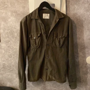 Olivgrön overshirt från Oscar Jacobson - Säljer en stilren olivgrön overshirt från Oscar Jacobson i storlek XS (passar även S). Skjortan har långa ärmar och knappar framtill samt två bröstfickor med knappstängning. Perfekt för en klassisk och tidlös look. Väldigt bra skick förutom defekt på bild 5 (Knappen kommer med så ni kan sy på själva! Därav pris) 