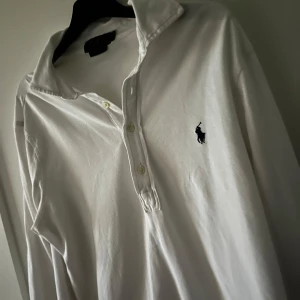 Vit pikétröja från Ralph Lauren - Säljer en klassisk vit pikétröja från Ralph Lauren med den ikoniska loggan på bröstet. Tröjan har långa ärmar och en knappslå med tre knappar. Perfekt för en stilren look.