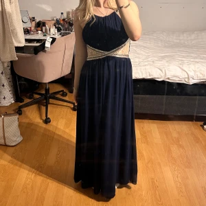 Mörkblå långklänning med spetsdetaljer - Elegant mörkblå långklänning med vackra spetsdetaljer i midjan. Klänningen har en halterneck-design och är ärmlös, vilket ger en stilren look. Perfekt för festliga tillfällen💘