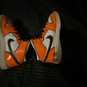 Nike sneakers i orange och vit - Snygga Nike sneakers i en fräck kombination av orange och vit med svarta detaljer. Skorna har en klassisk hög modell med snörning och den ikoniska Nike-loggan på sidan. Perfekta för dig som vill sticka ut med stil.