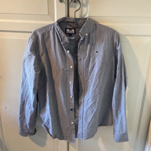 Grå skjorta från Weekend Offender - Säljer en stilren grå skjorta från Weekend Offender i storlek M. Skjortan är slim fit och tillverkad i 100% bomull. Den har långa ärmar och knappar framtill samt en liten broderad logga på bröstet.