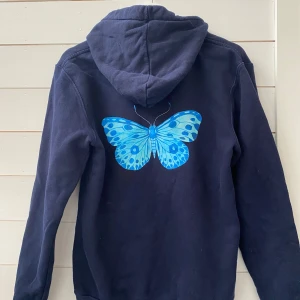Mörkblå hoodie med fjärilsmotiv från The Cool Elephant - Säljer en mörkblå hoodie från The Cool Elephant med en stor blå fjäril på ryggen och en mindre fjäril på bröstet. Tröjan har en klassisk känguruficka och justerbar huva med dragsko. Perfekt för en avslappnad stil.