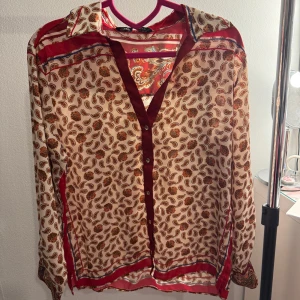 Mönstrad blus från Zara - Säljer en snygg blus från Zara med paisleymönster i rött och beige. Blusen har långa ärmar och knappar framtill. Perfekt för att ge en färgglad touch till din outfit!