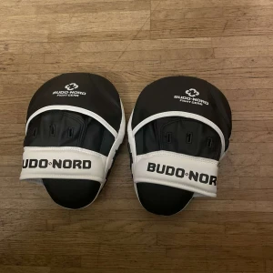  träningsmittsar Budo-Nord - Säljer ett par svarta och vita träningsmittsar från Budo-Nord. Perfekta för kampsportsträning med en bekväm passform och robust design. Mittsarna är justerbara remmar för säker användning.