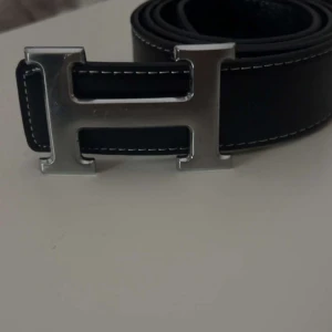 Svart bälte med silverfärgat H-spänne - Snyggt svart bälte med ett stort silverfärgat H-spänne. Bältet har vita sömmar som ger en fin kontrast. Perfekt för att ge en stilren touch till din outfit.