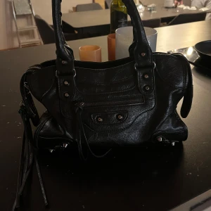 Svart handväska med dragkedja - Snygg svart handväska i skinn med dragkedja och nitar. Perfekt för att ge din outfit en fin touch. Väskan har dubbla handtag och en stilren design som passar till många tillfällen. Axelrems band tillkommer. Köpt för 1300kr. Skriv för fler bilder. Pris kan diskuteras 
