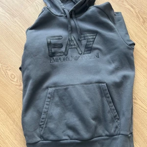 Grå hoodie från Emporio Armani - Säljer en stilren grå hoodie från Emporio Armani med EA7-logga på bröstet. Tröjan har en praktisk magficka och justerbar huva med dragsko. Perfekt för en avslappnad look. Den är som helt ny bara använd ett par grejer