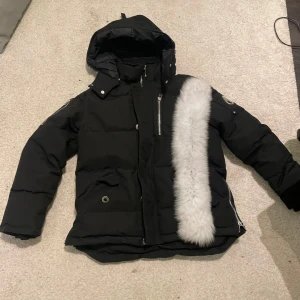 Svart dunjacka från Moose Knuckles - Säljer en svart dunjacka från Moose Knuckles med avtagbar huva och snygga detaljer. Jackan har en dragkedja framtill, flera fickor och en vit pälsdetalj på huvan. Perfekt för kalla dagar!