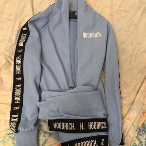 Ljusblå jacka från Hoodrich - Säljer en ljusblå jacka från Hoodrich med dragkedja och svarta detaljer längs ärmarna. Jackan har texten 'Hoodrich' tryckt i vitt på bröstet och längs ärmarna. Perfekt för en sportig look!