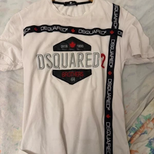 Vit t-shirt från Dsquared2 - Snygg vit t-shirt från Dsquared2 med svart och röd logotypdesign. Den har korta ärmar och ett unikt bandmönster med texten 'DSQUARED2' och röda lönnlöv. Perfekt för en stilren look.