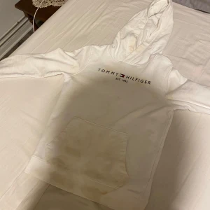 Vit hoodie från Tommy Hilfiger - Säljer en vit hoodie från Tommy Hilfiger med klassisk logga på bröstet. Hoodien har en stor ficka framtill och en bekväm huva. Perfekt för en avslappnad stil.