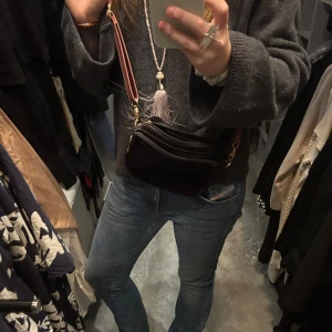 Svart axelväska med randigt band - Snygg svart axelväska med ett randigt axelband i rött och beige. Väskan har flera fack med dragkedjor och guldiga detaljer. Perfekt för att bära med sig det viktigaste med stil.