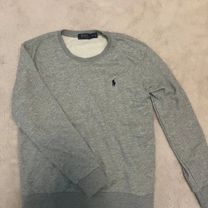 Grå sweatshirt från Polo Ralph Lauren - Klassisk crewneck i en stilren ljusgrå nyans, tillverkad i mjuk och bekväm bomull. Designad med ribbade muddar och broderad logga på bröstet för en exklusiv touch. Ett perfekt basplagg som kombinerar komfort och elegans.