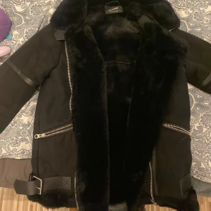 Svart jacka från Pull&Bear - Snygg svart jacka från Pull&Bear med pälsfoder och dragkedjor. Jackan har bältesdetaljer och en mysig krage. Har endast används ett fåtal gånger och är i mycket bra skick. 