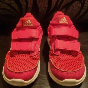 Rosa träningsskor barn från Adidas - Säljer ett par rosa träningsskor barn från Adidas med två kardborreband för enkel justering. Skorna har en vit sula och de klassiska tre ränderna i vitt på båda sidorna. Ovandelen är i ett andningsbart meshmaterial. Mycket fint skick, med väldigt få märken,