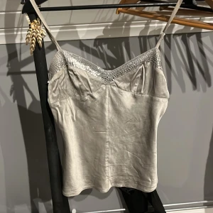 Silvrig satinlinne med paljettdetaljer - Snygg silvrig linne i satin med smala axelband och glittriga paljettdetaljer längs urringningen. Perfekt för en kväll ute eller en speciell tillställning. Enkel och elegant design som ger en lyxig känsla. AI