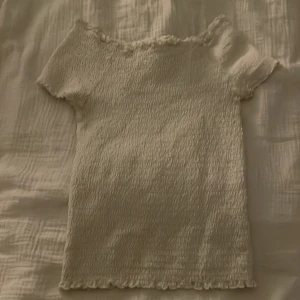 Vit ribbad topp - Säljer en söt vit ribbad topp med korta ärmar och volangdetaljer vid hals och nederkant. Perfekt för en avslappnad stil. Passar storlek 146/152.