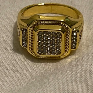 Guldfärgad ring med glittriga stenar - Säljer en snygg guldfärgad ring med en fyrkantig design på toppen, dekorerad med massor av glittriga stenar. Ringen har också ett coolt mönster på sidorna som ger den en unik look. Perfekt för att blinga upp vilken outfit som helst! NY storlek 8