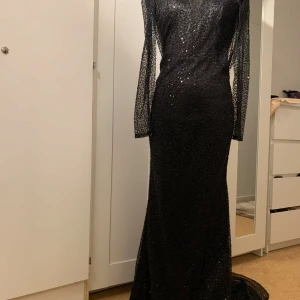 Svart glittrig långklänning - Säljer en elegant svart långklänning med glittrande detaljer. Klänningen har långa ärmar och en vacker öppen rygg som ger en sofistikerad look. Perfekt för festliga tillfällen där du vill glänsa!