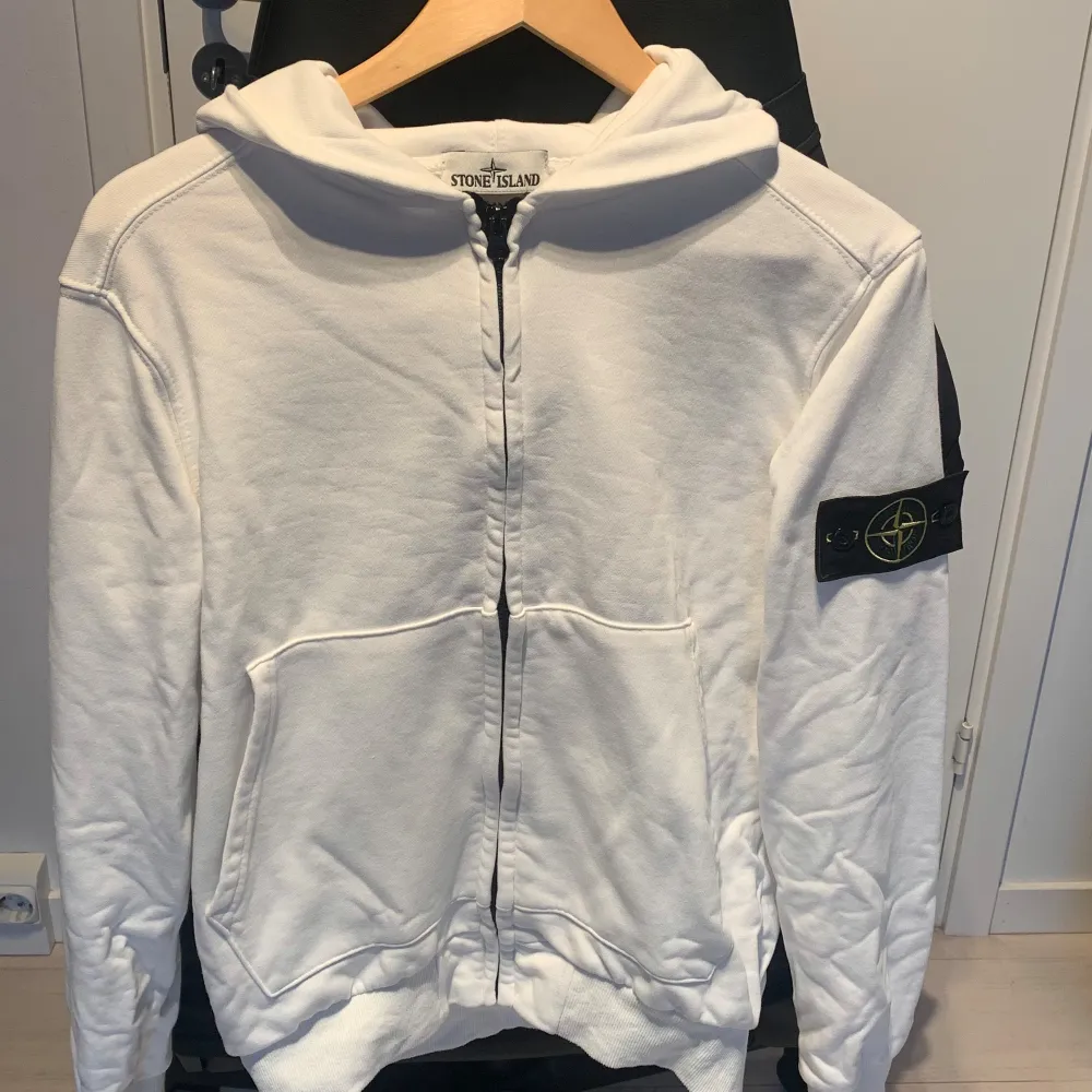 Säljer en snygg vit hoodie från Stone Island. Den har en dragkedja framtill och en ikonisk patch på ärmen. Perfekt för en stilren och avslappnad look. Passar bra till både vardag och fest!. Tröjor & Koftor.