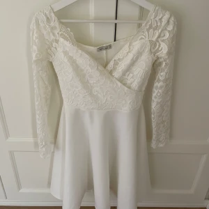Vit spetsklänning från NLY One - Säljer en superfin vit klänning från NLY One i storlek XS. Klänningen har ett vackert spetsmönster och långa ärmar. Den har en omlott-design framtill som ger en elegant touch. Perfekt för sommarens fester eller kanske till studenten! 💫