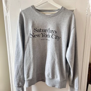 Grå sweatshirt från Saturdays New York City - Säljer en stilren grå sweatshirt från Saturdays New York City. Den har en klassisk rund hals och långa ärmar. Perfekt för en avslappnad look, oavsett om du hänger hemma eller är ute på stan. Texten på framsidan ger den en cool touch. 
