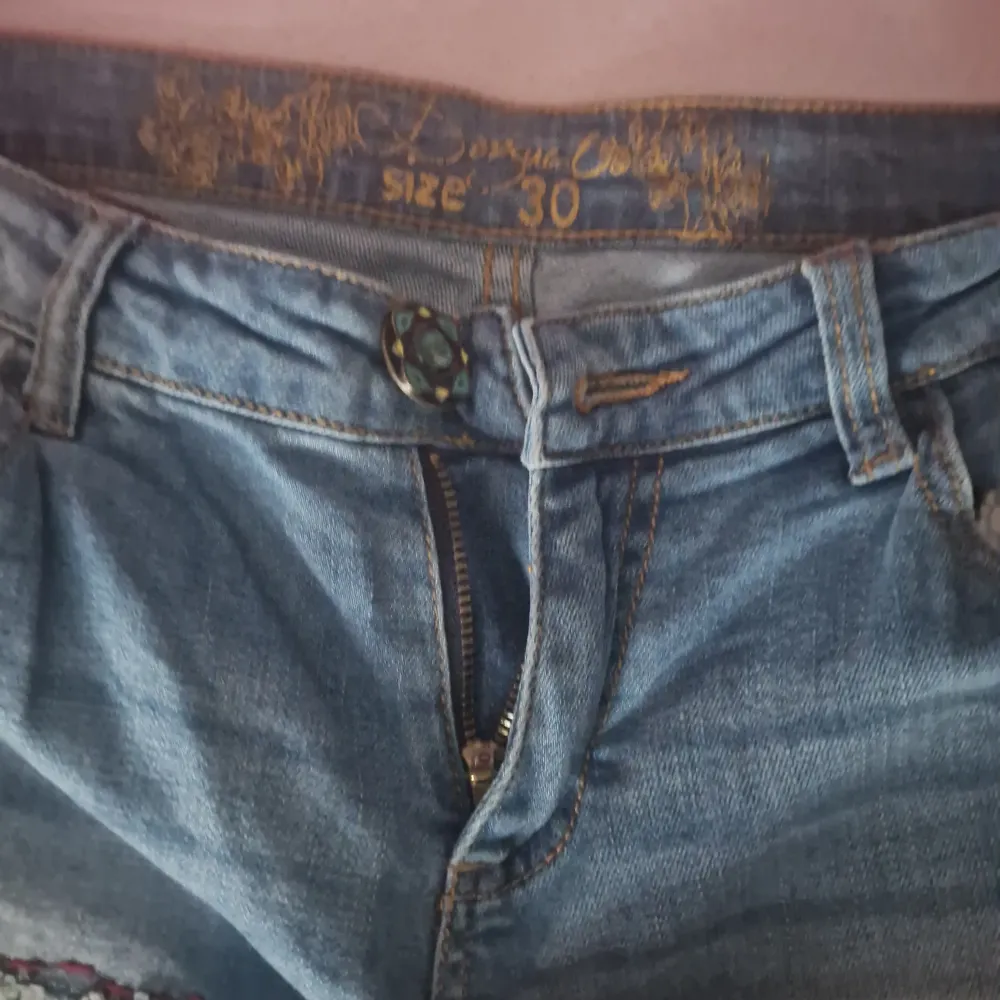 Säljer ett par coola jeans från Desigual i storlek 30. De är blå med unika broderade detaljer i färgglada mönster på fickorna. Jeansen har en sliten look med patchar och är perfekta för en avslappnad stil. De har en knapp och dragkedja framtill.. Jeans & Byxor.