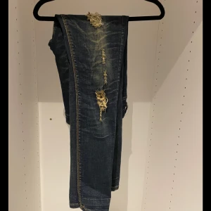 Blå slitna jeans - Säljer ett par blå jeans med coola slitningar och en unik insida med mönster. Jeansen har en klassisk blå färg och är i en välanvänd stil med slitna detaljer som ger dem en edgy look. Insidan av byxorna har ett rutigt och mönstrat tyg i beige och brunt, vilket gör dem extra speciella.