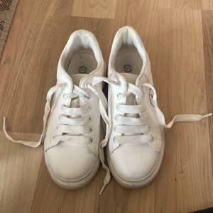 Vita sneakers med Doraemon-motiv - Säljer ett par vita sneakers med ett coolt Doraemon-motiv på sidan. Skorna har snörning och är i syntetmaterial. Perfekta för en avslappnad stil och passar till nästan allt. Använda men i bra skick!