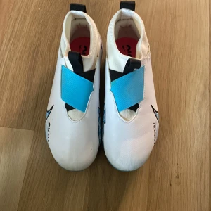 Vita och blå fotbollsskor från Nike - Säljer ett par snygga fotbollsskor från Nike i vitt och blått med coola detaljer. Skorna har en modern design med snörning och en unik mönstrad sula i blått och rosa. Perfekta för fotbollsträning eller match!