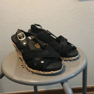 Svarta sandaler med kilklack från La Redoute - Säljer ett par snygga svarta sandaler från La Redoute i storlek 36. De har en bekväm kilklack med flätad detalj i naturfärg och justerbart spänne. Perfekta för sommaren och passar både till vardags och fest!