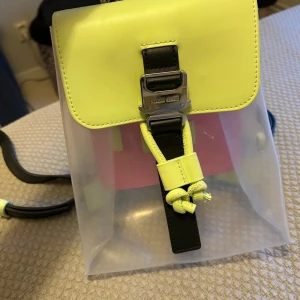 Transparent ryggsäck med neongula detaljer - Säljer en supercool transparent ryggsäck från Tommy Hilfiger med neongula detaljer. Den har en unik design med en svart spänne framtill och justerbara svarta axelremmar. Perfekt för att ge din outfit en färgklick! 🖤💛 Nyskick 