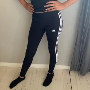Svarta leggings från Adidas - Säljer ett par svarta leggings från Adidas med de klassiska vita ränderna längs sidorna. De är högmidjade och har en tight passform, perfekt för träning eller en avslappnad dag. Materialet är mjukt och stretchigt för bästa komfort.