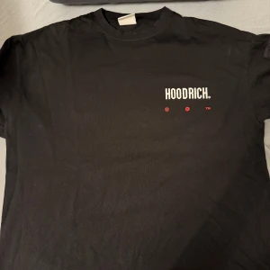 Svart t-shirt från Hoodrich - Snygg svart t-shirt från Hoodrich med tryck både fram och bak. T-shirten har en liten ficka med dragkedja på ärmen och texten 'Hoodrich' i vitt och rött. Perfekt för en casual look.