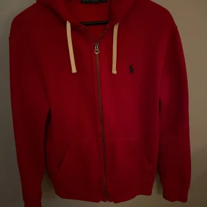 Röd hoodie från Polo Ralph Lauren - Säljer en snygg röd hoodie från Polo Ralph Lauren med dragkedja och justerbara snören i huvan. Den har en broderad logga på bröstet och praktiska fickor framtill. Perfekt för en avslappnad stil. Väldigt bra skick och använd några gånger.
