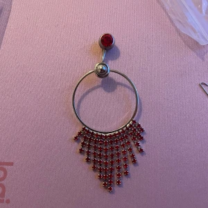 Navelpersing med röd sten och fransdetaljer - Snyggt smycke med en stor röd sten och hängande fransdetaljer i rött. Perfekt för att ge en extra touch till din stil.