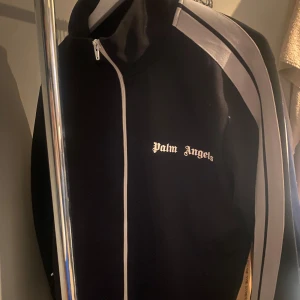 Svart track jacket från Palm Angels - Snygg svart track jacket från Palm Angels med vita ränder längs ärmarna och dragkedja framtill. Jackan har en sportig look med broderad logga på bröstet. Perfekt för en casual stil.