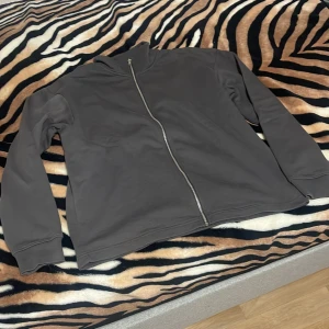 Grå huvtröja med dragkedja - Säljer en stilren grå huvtröja med dragkedja. Tröjan har långa ärmar och en bekväm passform, perfekt för vardagsbruk. Den är tillverkad i ett mjukt material som ger en skön känsla.