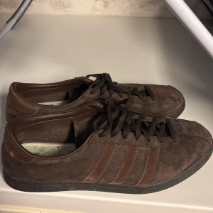 Tobaksbruna adidas  - Säljer ett par adidas tobacco sneakers från Adidas i mocka med klassiska ränder på sidan. Skorna har snörning och en bekväm innersula med Adidas-logga. Perfekta för en avslappnad stil.