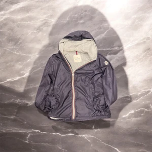 Mörkblå vindjacka från Moncler - Snygg mörkblå vindjacka från Moncler med huva och dragkedja i regnbågsfärger. Jackan har ett broderat Moncler-märke på ärmen och en grå insida. Perfekt för blåsiga dagar! Det är dock ett litet hål innanför som man inte märker. Äkta ✅