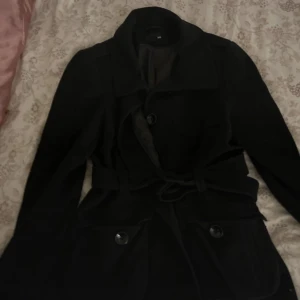 Svart kappa från H&M - Snygg svart kappa från H&M med knappar och knytband i midjan. Perfekt för kyligare dagar och ger en stilren look. Passar bra till både vardag och mer uppklädda tillfällen.