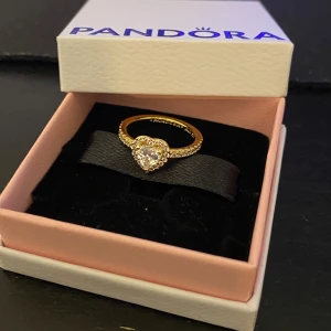 Guldfärgad ring med hjärta från Pandora - Elegant guldfärgad ring från Pandora med ett glittrande hjärta i mitten. Äkta 14k guldpläterat. Säljer denna ring eftersom jag råka köpa fel storlek till min tjej det är storlek 48 i diameter. Perfekt att ge sin tjej. Mitt Pris 800kr, original pris 1200kr