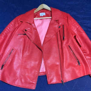 Röd skinnjacka från Zizzi - Snygg röd skinnjacka från Zizzi med dragkedjor och en cool bikerstil. Jackan har långa ärmar och flera praktiska fickor med dragkedja. Perfekt för att ge din outfit en edgy touch.