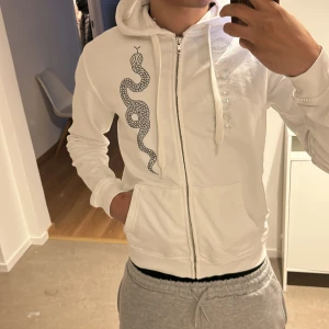 Vit hoodie med ormmönster från Trapper - Säljer en vit hoodie från Madebytrappers med dragkedja och ett coolt ormmönster på framsidan. Perfekt för en avslappnad stil. Den har en bekväm passform och huva. Pris kan diskuteras!